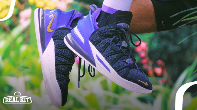 A person in white socks wearing a pair of black LeBron 18s featuring purple heels, gold Swooshes, and white midsoles.
