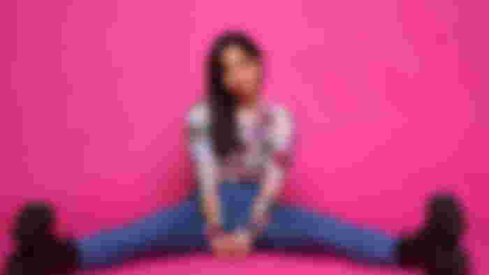 Kayane sitting down and looking to the side with a bright pink background