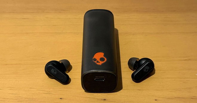 A pair of black Skullcandy-branded wireless earbuds either side of their charging case which features the Skullcandy logo in orange on the front.