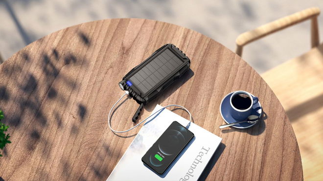 A rugged, black power bank being used to charge a black phone while sat on a brown wooden table with a blue teacup.