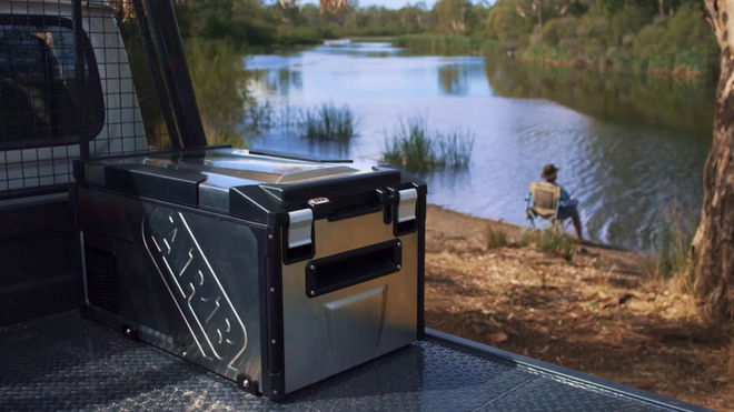 A silver and black portable fridge sat on the back of a vehicle in front of a lake.