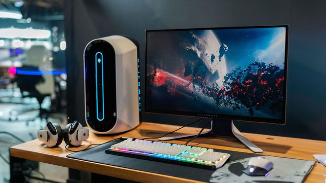 A white and black monitor sat on a brown wooden desk next to a white PC, headset, backlit keyboard, and mouse.