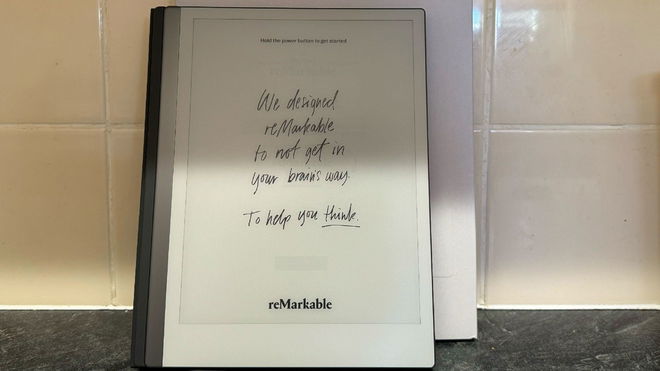 The reMarkable 2 device standing in front of its box and in front of a tiled wall.