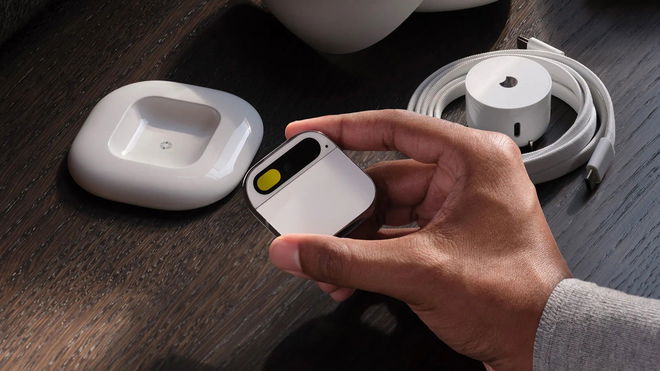A person holding the Humane AI Pin in their hand in front of the charging block and the charging case