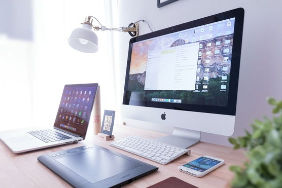 a desk with a computer , laptop , tablet , phone and lamp .
