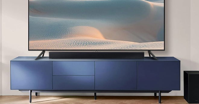 A long, black, rectangular soundbar sat on a blue TV stand with a flatscreen TV behind it.