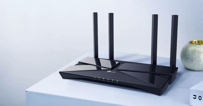 A black WiFi router with four antennae sat on a white table top next to a vase.