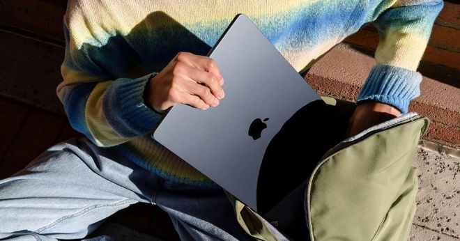 Someone in a blue and yellow jumper putting a Midnight-coloured Apple MacBook into a green rucksack.