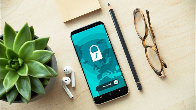 A black smartphone with a VPN lock on the display sat next to a plant, AirPods, a pencil, and a pair of glasses.