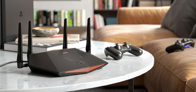 A black WiFi router with red trim at the front and back sat on a white table next to a black PlayStation controller.