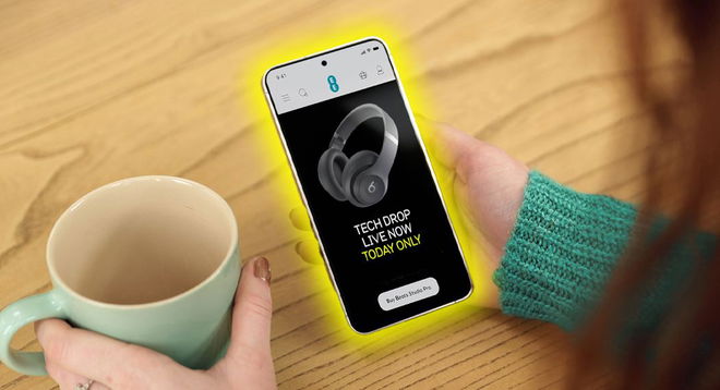 Someone in a green jumper holding a mug in one hand and a phone in the other. On the phone, an EE Tech Drop on a pair of Beats headphones.
