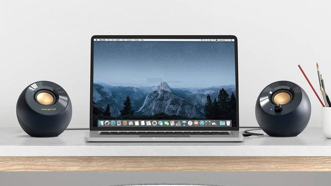 A set of two small, round, and black speakers either side of a laptop sat at a desk.