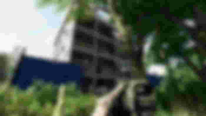 A first-person screenshot of a player holding their rifle at the ready while looking upwards at a large building under construction through some trees.