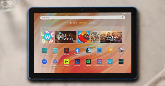 A black and light blue Amazon tablet with the home screen in front of an orange background on the display.