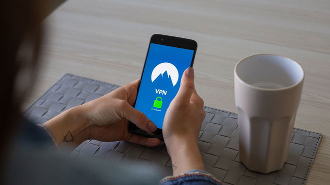 Someone holding a black phone with a blue VPN service loaded up on the screen next to a white cup. 