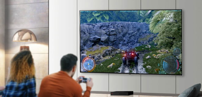 Two people playing a driving video game on a large flatscreen TV mounted to a white wall.