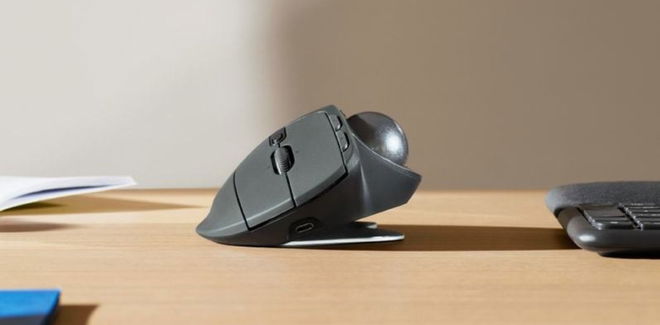 A graphite-coloured ergonomic mouse with a trackball on the left side sat on a light brown desk.