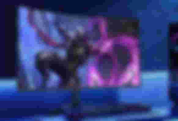 A black gaming monitor sat on a desk in a blue-lit room, the monitor featuring a armoured video game character with pink energy coming from its arm on the display.