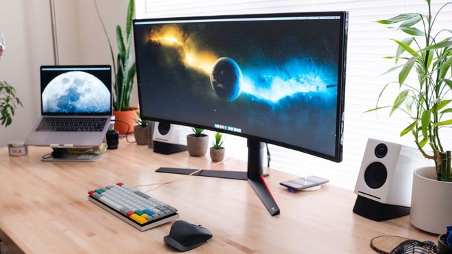 A curved black monitor sat on a wooden desk with an old-school grey keyboard, mouse, laptop, and speakers.